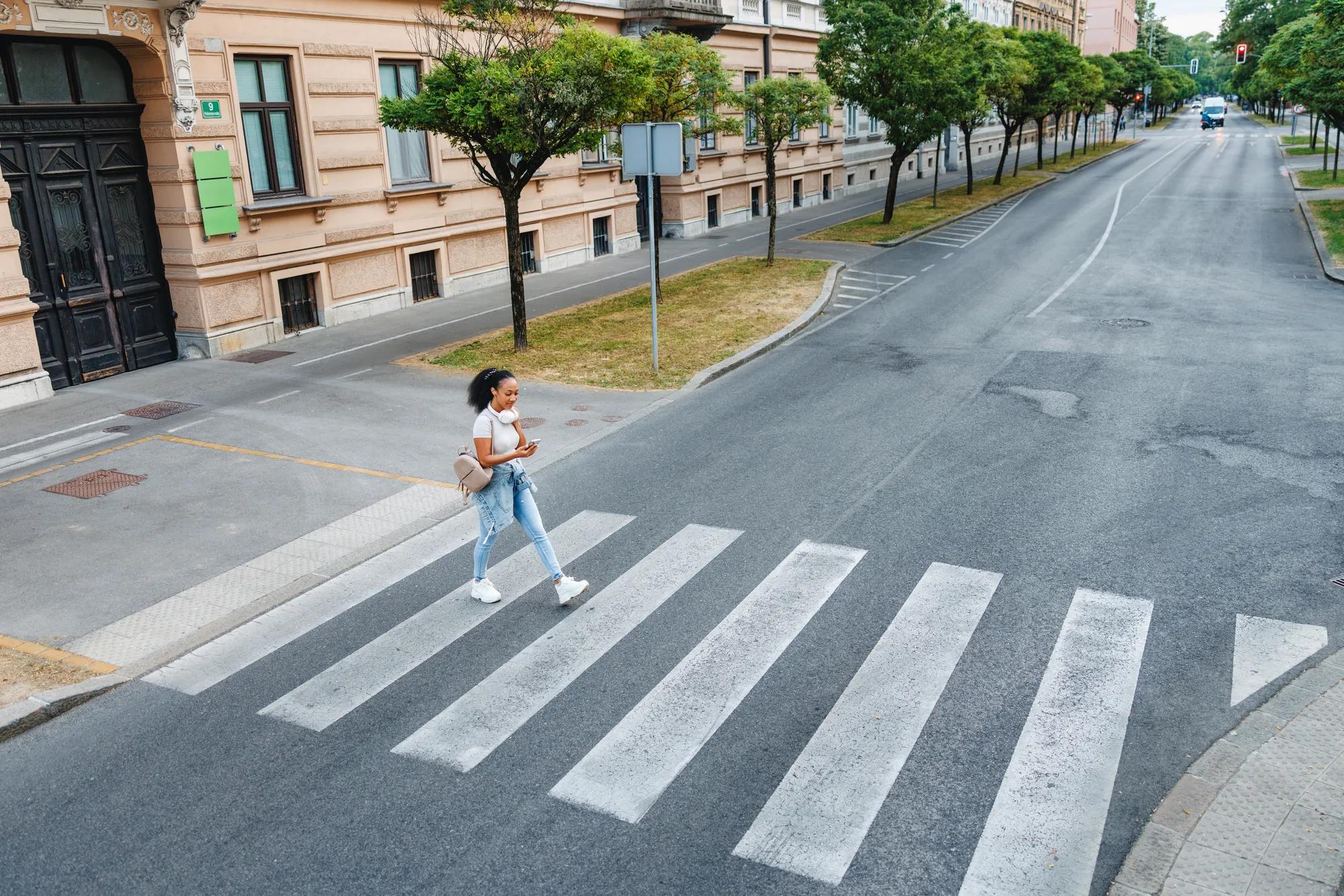 A young adult crosses a city street on a crosswalk, focused on her phone. The scene reflects daily urban life, independence, and technology use in a bustling metropolitan area. Captured from a high angle.