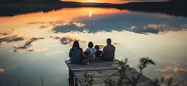 family of four people sitting on the shore on a wooden bridge of a large lake in summer and watching the beautiful sunset with dog back view